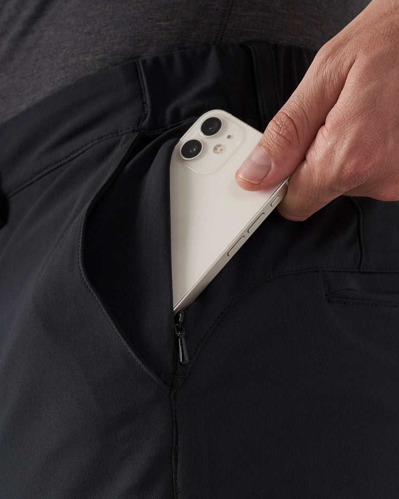 A close up detailed shot of a hand sliding a white mobile phone into a front zippered pocket to illustrate the size and depth of the pocket.