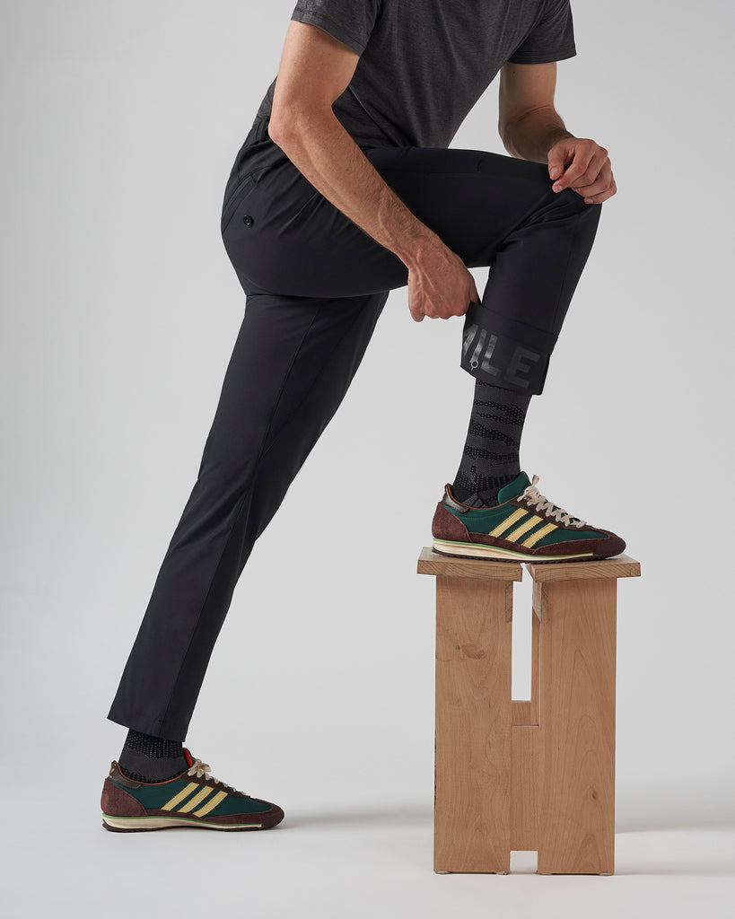 The male model is lunging with a foot up on a raised stool, showcasing the four-way stretch and flexibility of the trousers he is wearing. He has folded up the trouser cuff to showcase the reflective inner panel, and pairs the black commuter trousers with wales bonner x adidas sneakers and a grey t-shirt