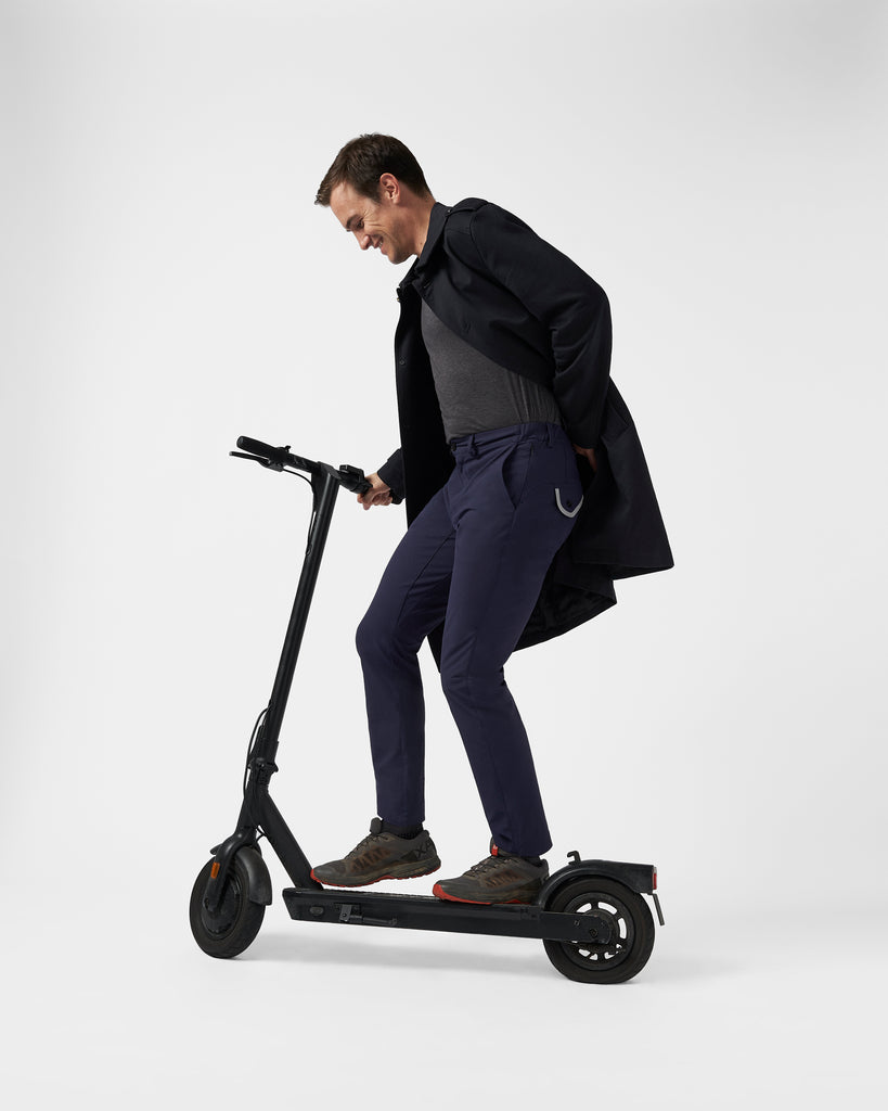 A man rides a black e-scooter (electric scooter) wearing the navy commuter trousers, paired with grey salomon shoes and a grey long coat. | The Navy Ones | Performance-Wear Brand | Buy Performance-Wear M1LE