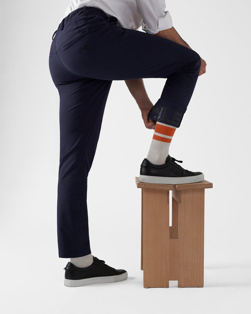 Male model illustrating the flexibility of the trousers by stepping up onto a wooden stool with one leg in an exaggerated fashion, highlighting the inner reflective trouser cuff at the same time. The navy trousers are paired with a white shirt and black leather sneakers.