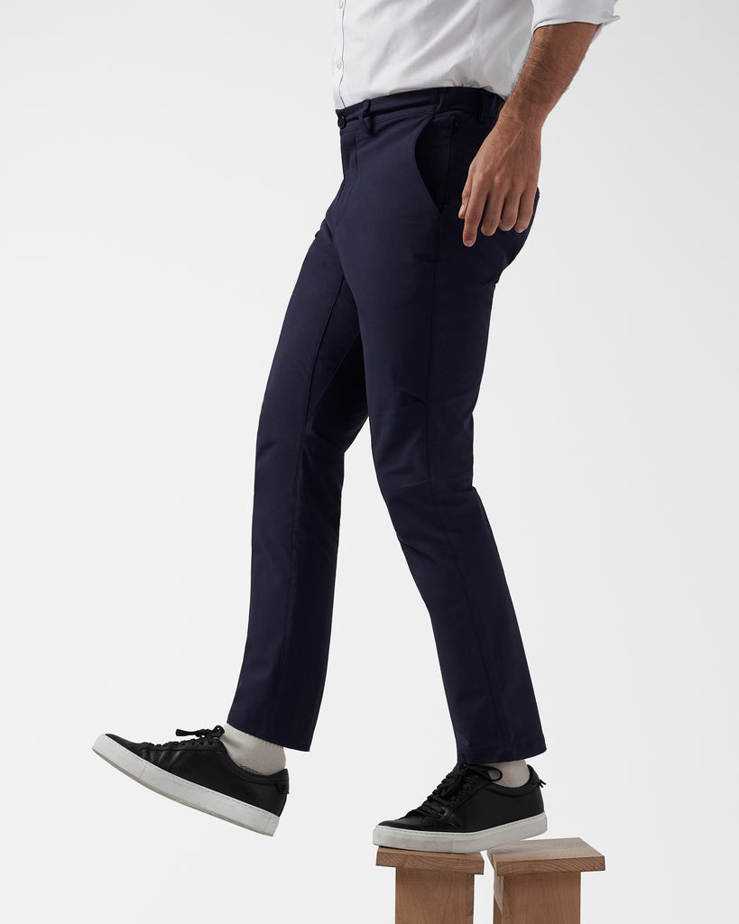 The model is stepping off of a wooden stool wearing navy workwear commuter trousers to illustrate their silhouette from a side view. The trousers are paired with a white office shirt and black leather sneakers.
