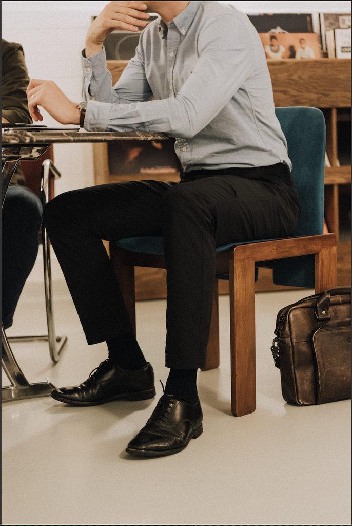 A man sits at an office desk wearing black M1LE trousers paired with a pale blue ralph lauren shirt, black church's shoes and a leather briefcase