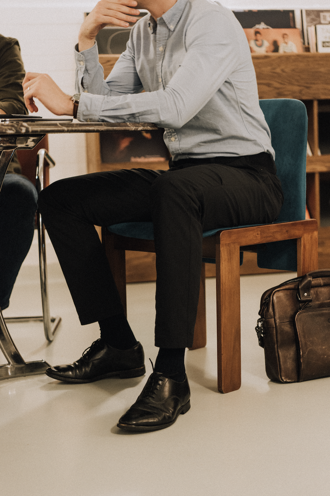 A man sits at an office desk wearing black M1LE trousers paired with a pale blue ralph lauren shirt, black church's shoes and a leather briefcase