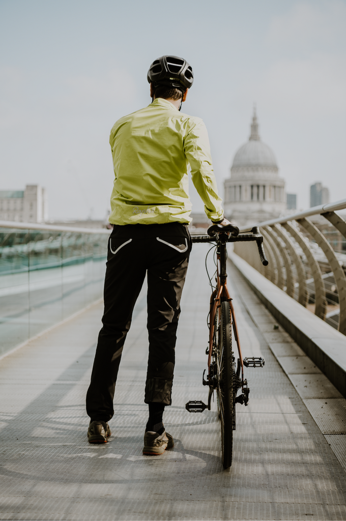 A man stands on the millennium bridge in london facing st.paul's cathedral wearing a luminous yellow cycling jacket, holding a genesis bicycle and wearing M1LE trousers with salomon shoes.